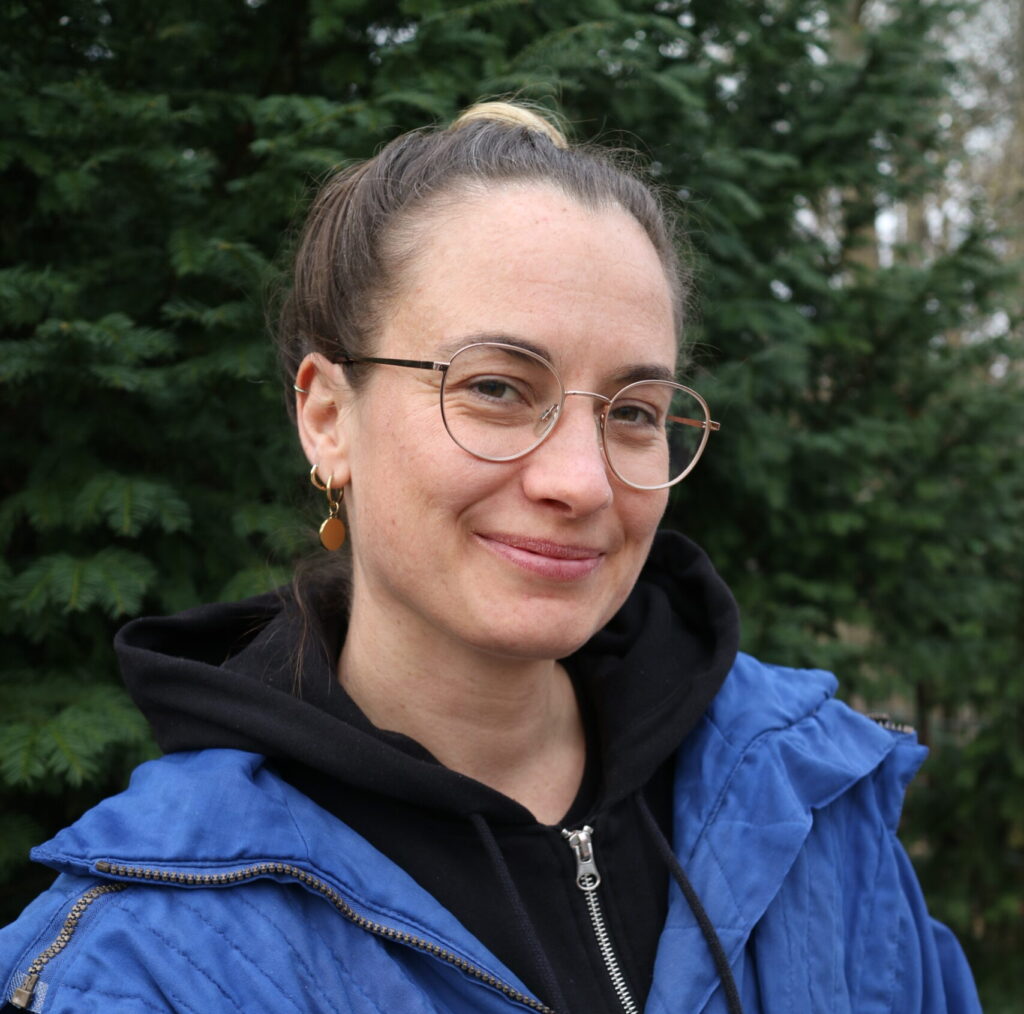 Bild einer etwa 30 jährigen Person mit dunkelbrauen zum Zopf gebundenen Haaren und Brille vor einem grünen Nadelbaum.